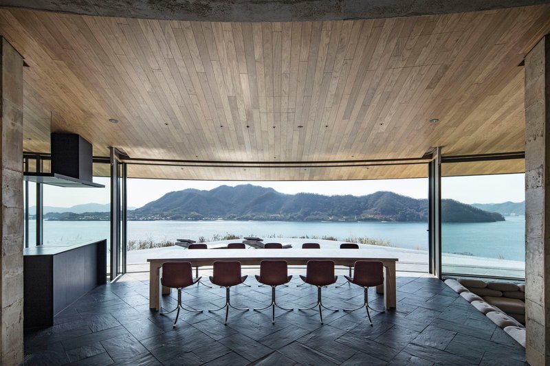Interior dining space with curved timber ceiling, floor-to-ceiling glazing and long table overlooking the water