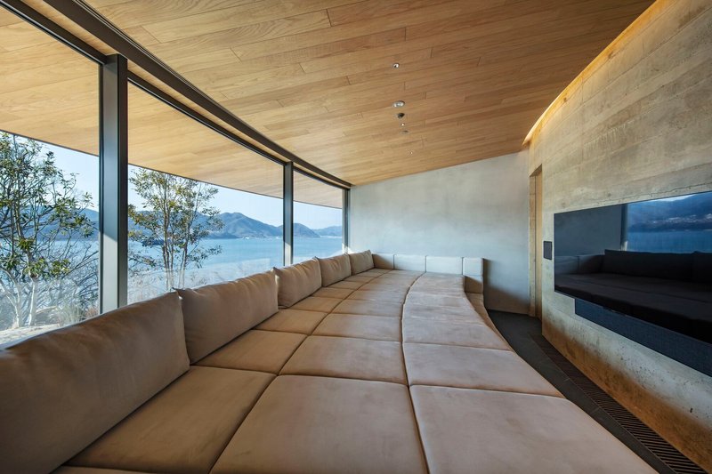 Curved lounge area with upholstered seating, concrete walls and continuous glazing framing the lake view