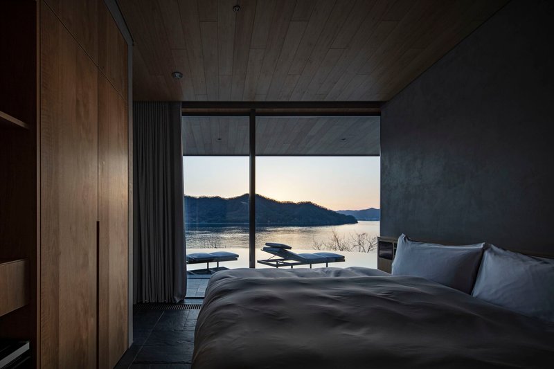 Bedroom with timber ceiling and concrete wall framing a lakeside view at dusk