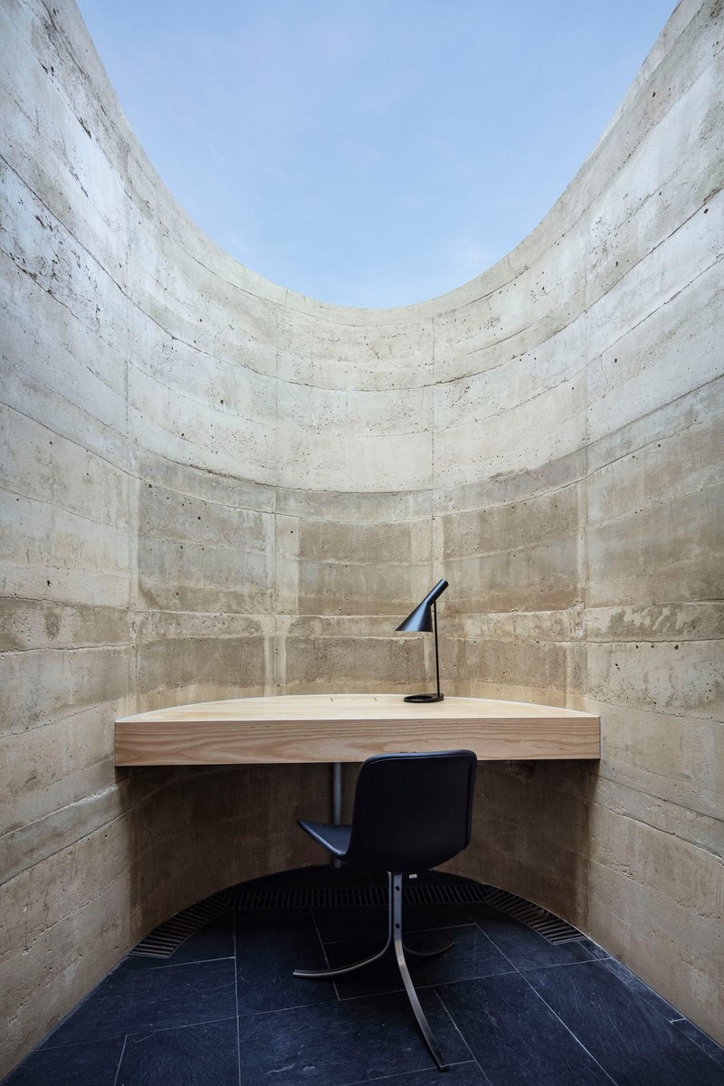 Small study alcove with board-formed concrete walls, timber desk and black swivel chair beneath an open skylight