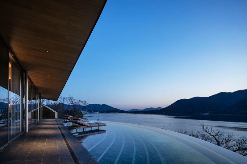 Lakeside infinity pool and glazed colonnade beneath a curved timber soffit at dusk