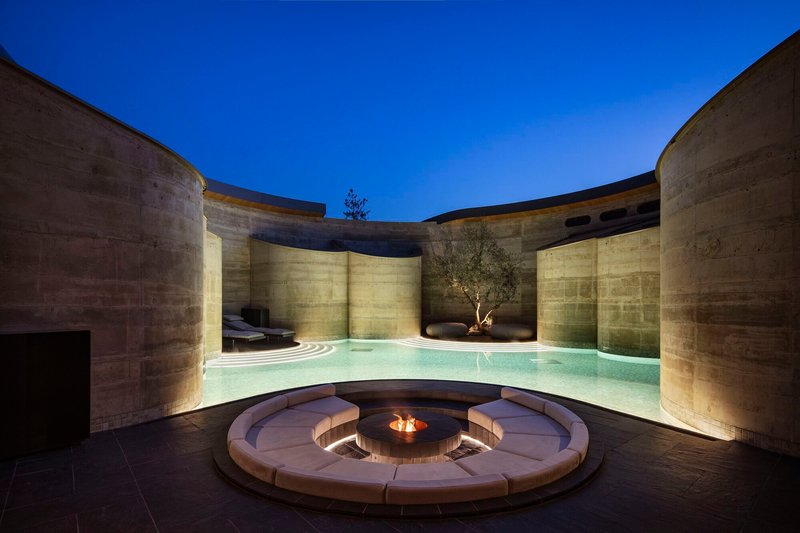 Curved swimming pool and circular fire pit seating area enclosed by uplighting concrete walls at night