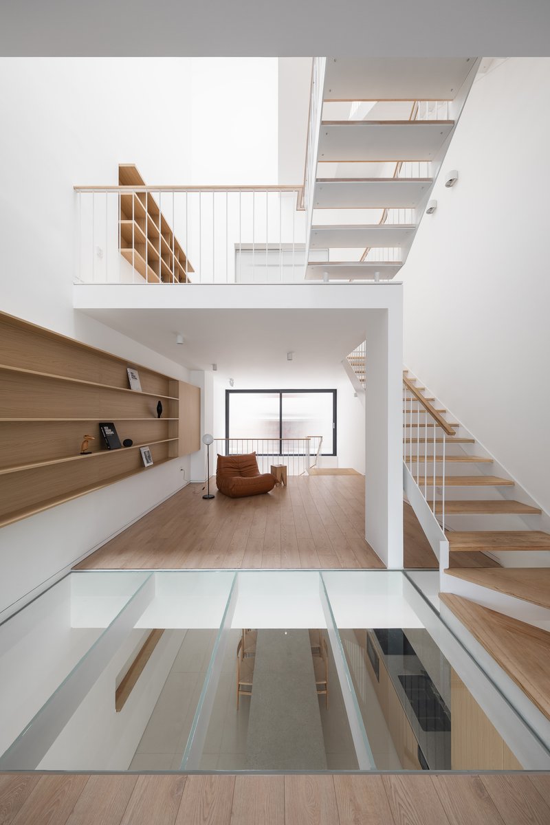 Double-height living space with timber stairs, glass floor panels, and built-in shelving along white walls