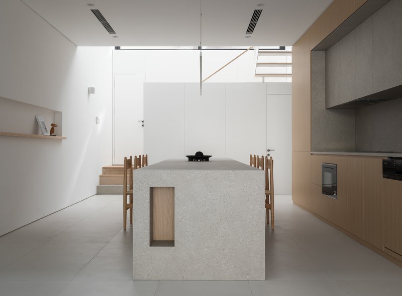 Open kitchen with terrazzo island and timber dining chairs beneath a floating upper stair