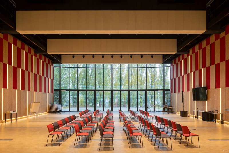 Assembly hall with red-and-yellow acoustic panels and rows of chairs facing a glazed forest view
