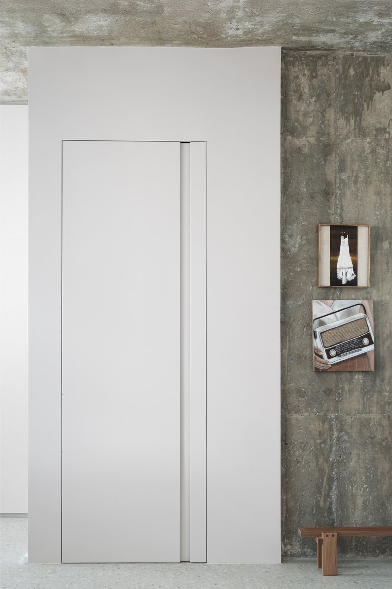 Flush white pocket door beside exposed concrete wall with wooden shelves and a vintage radio