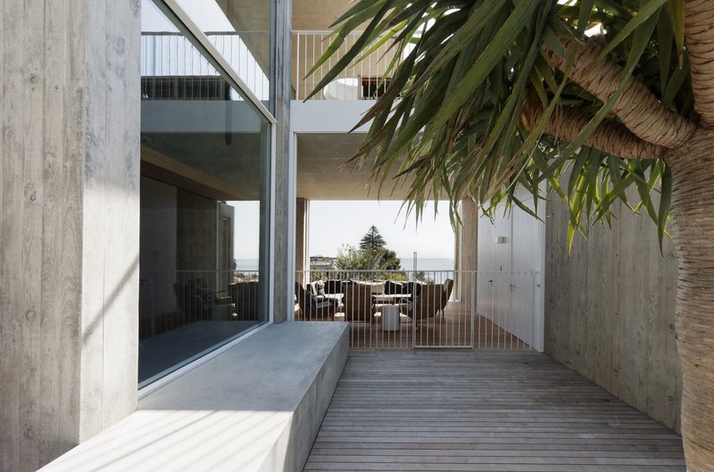 Timber deck passage beneath a concrete soffit with palm fronds overhead and sea view beyond