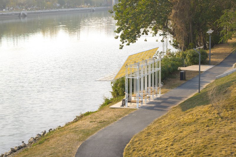 Timber and white steel pavilion with translucent roof panels along a riverside path at sunset