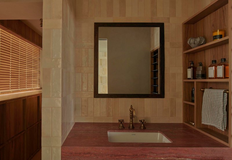Bathroom vanity with red stone countertop, framed mirror and timber shelving beside a slatted timber blind