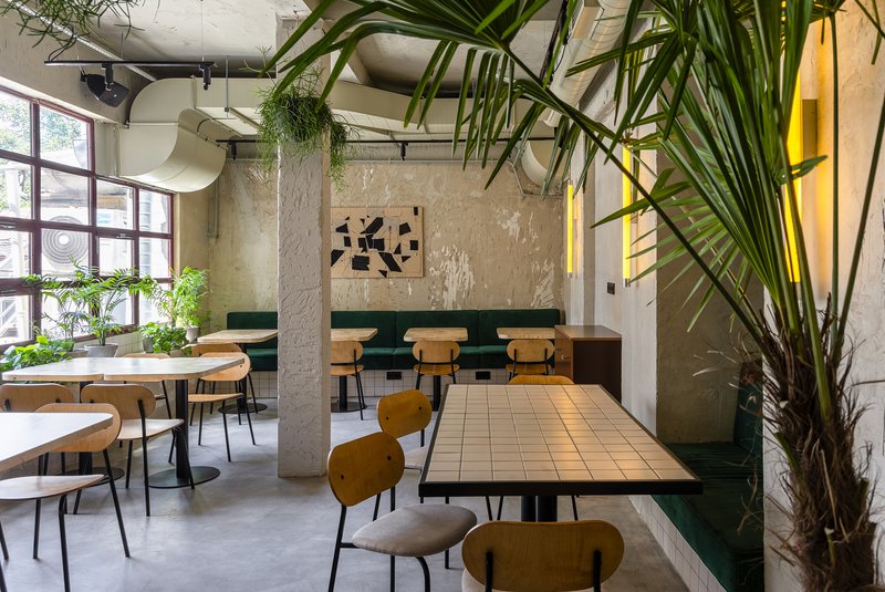 Dining area with exposed concrete ceiling, large palms, weathered plaster walls and tile-topped tables under natural light