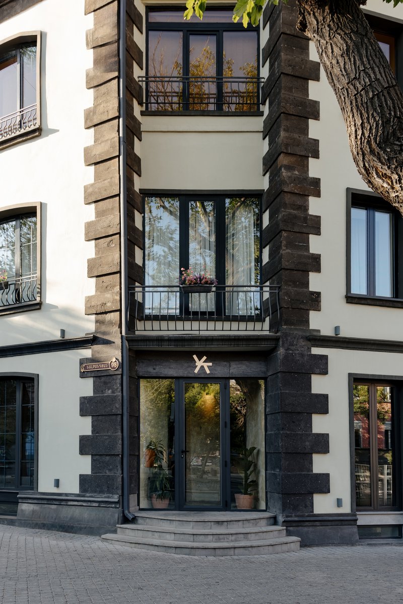 Street facade with white plaster and dark stone quoins framing balconies and glazed entry beneath a tree