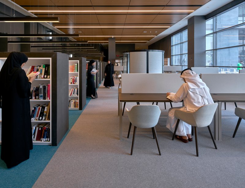 Library reading area with grey shelving and tables where visitors browse books beneath linear ceiling lights