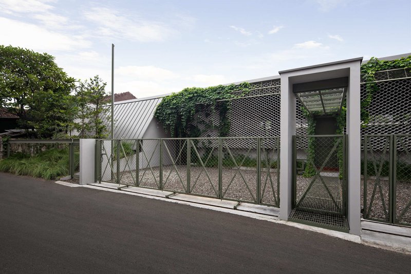 Street view of perforated metal fence with gate opening and climbing vines under a partly cloudy sky