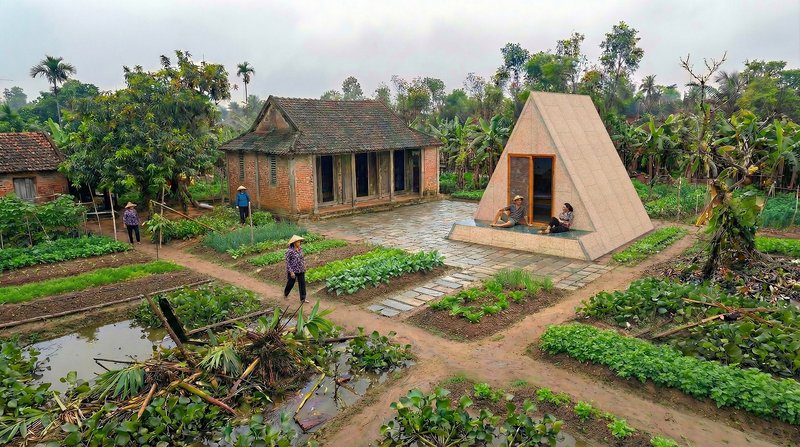 Triangular shelter on a concrete platform in a rural garden with vegetable plots and traditional houses