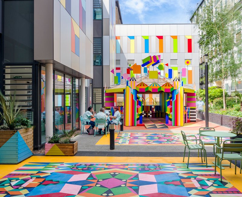 Courtyard with geometric paving and rainbow-striped pavilion entrance framed by residential buildings and diners at tables