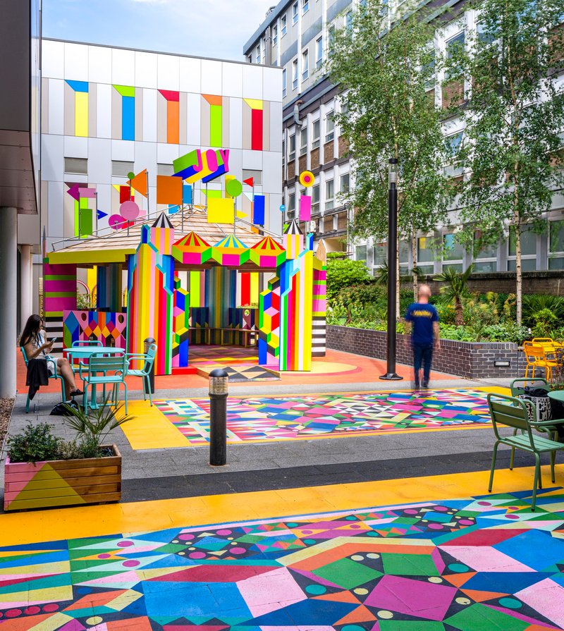 Outdoor pavilion with striped multicolor facade and matching ground graphics beside planted beds and seating