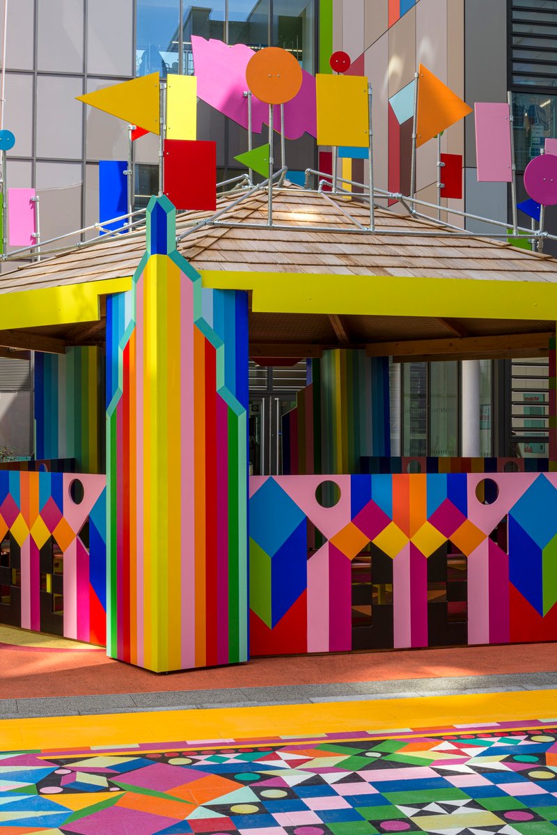 Pavilion structure with painted vertical stripe columns supporting a slatted timber roof under cloudy sky
