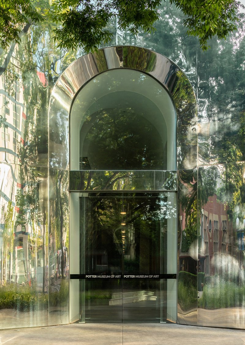 Polished metal arch and glass doorway reflecting foliage and adjacent buildings in afternoon light