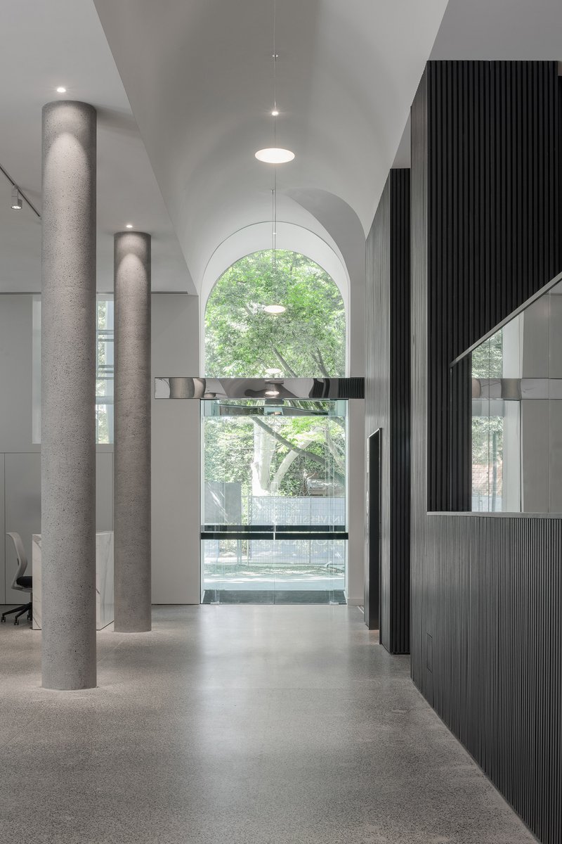 View through vaulted corridor with circular concrete columns toward arched window framing green foliage outside