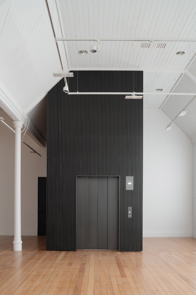 Black ribbed elevator core set against white paneled ceiling and timber floor in gallery hall
