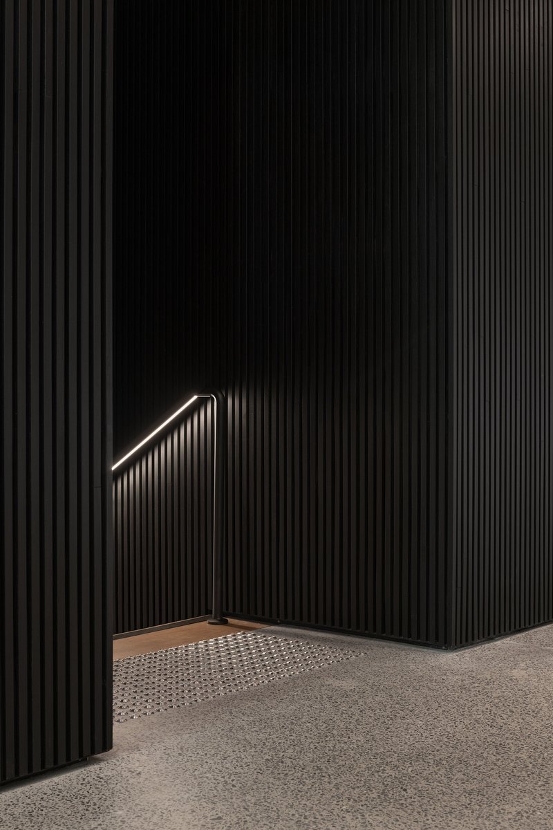 Black vertically-fluted wall with illuminated triangular handrail above a recessed perforated floor panel