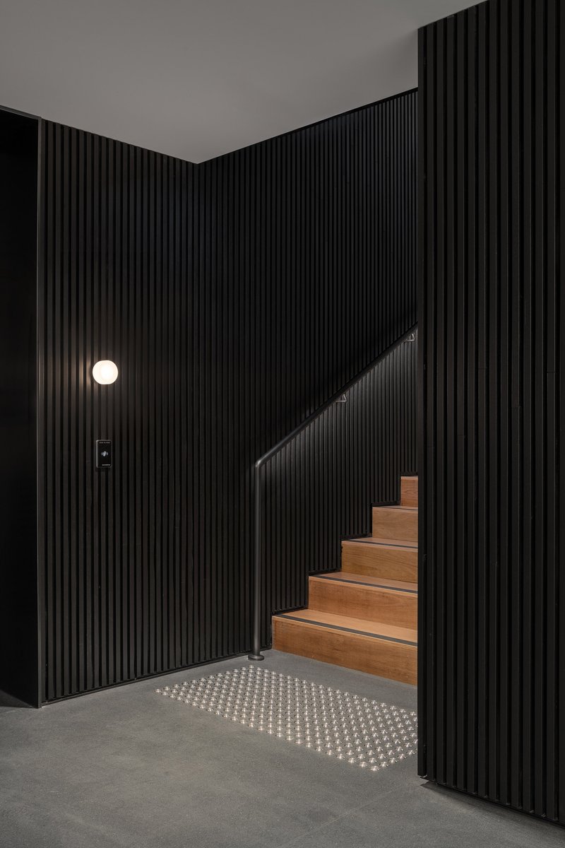 Timber staircase rising between black fluted walls with a circular wall sconce and illuminated floor grille