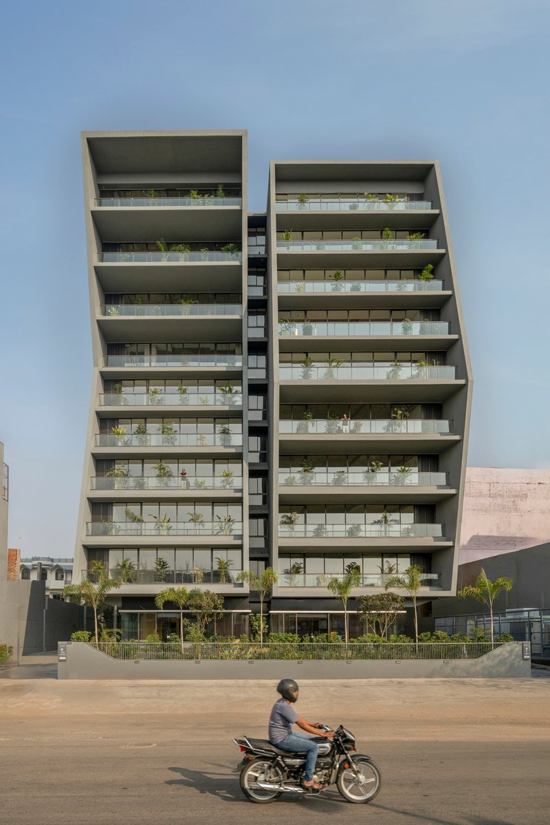 Street view of twin residential towers with deep concrete balconies and planted terraces, motorcyclist passing in foreground