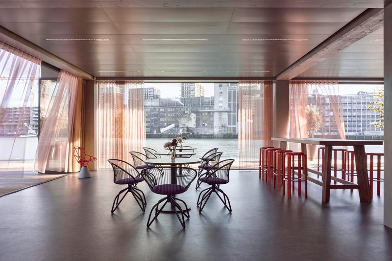 Dining area with metal frame chairs and floor-to-ceiling glazing overlooking a waterfront cityscape