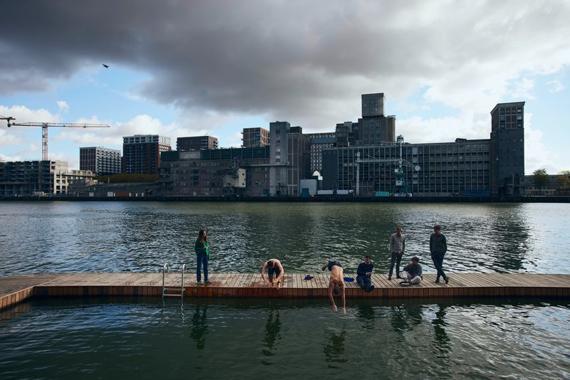 Timber swimming dock with people and a dog facing the converted industrial waterfront under dramatic clouds