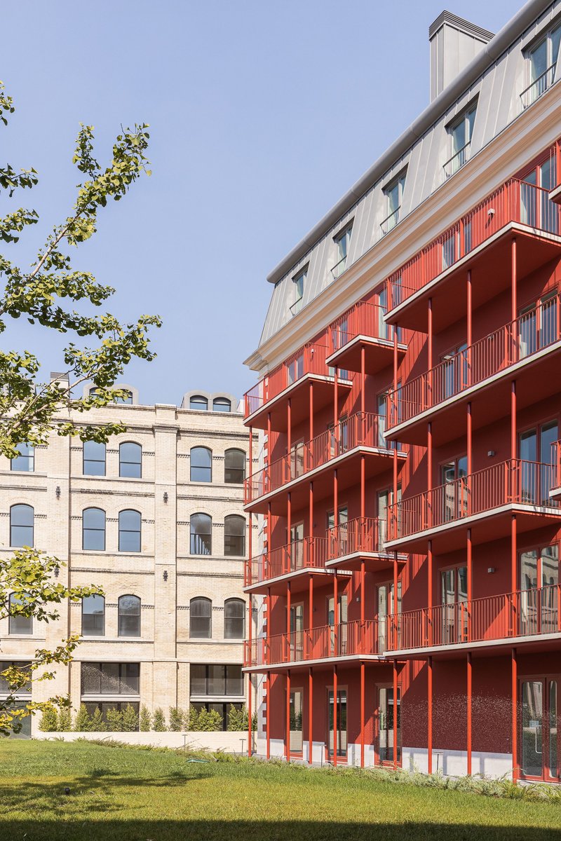 Red metal balconies stacked on a new block facing a historic arched brick building across a lawn