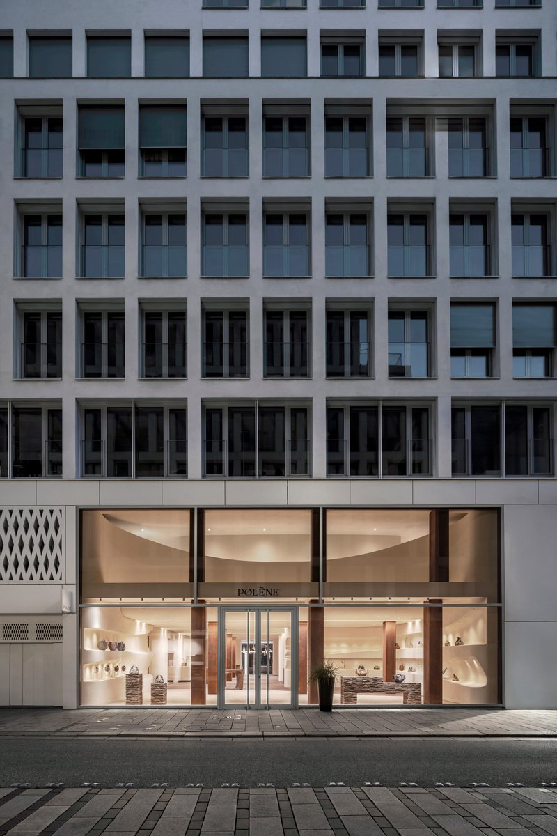 Street-level storefront with curved glass facade illuminated at dusk beneath a grey office tower