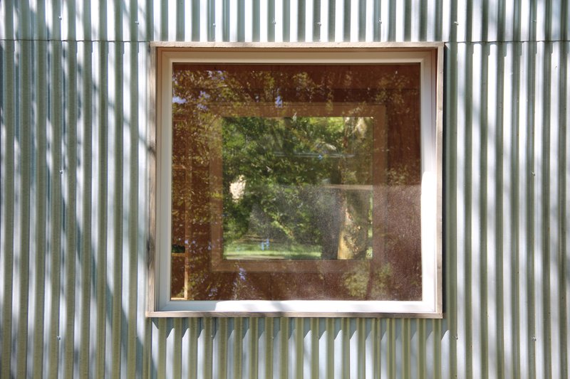 Square window with timber reveal set into corrugated metal cladding reflecting the wooded surroundings