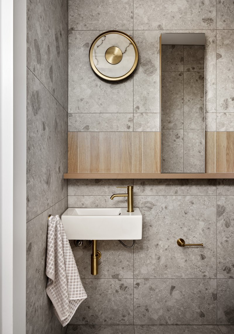 Wall-mounted basin with brass fittings below a timber and mirror cabinet on grey tile