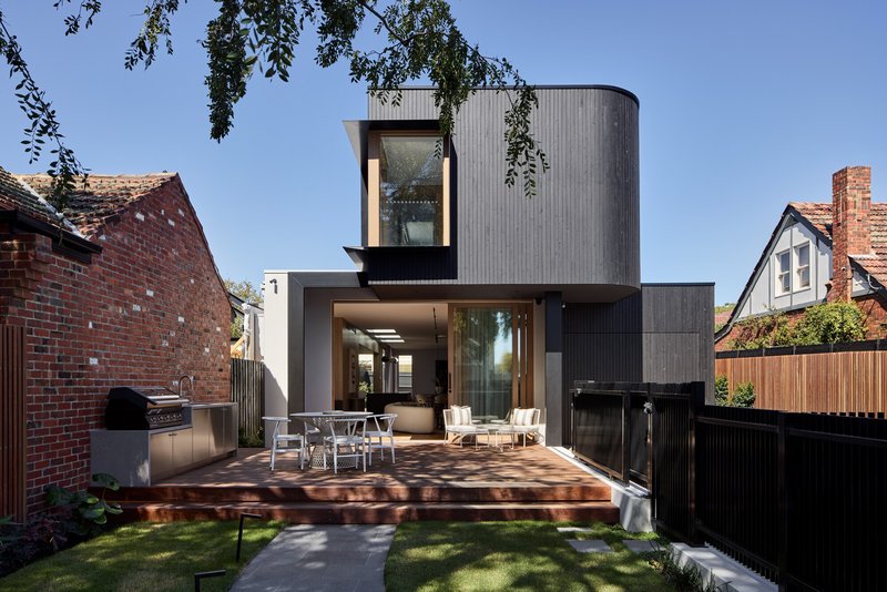 Rear garden view showing timber deck with outdoor dining and cylindrical upper volume clad in grey ribbed panels