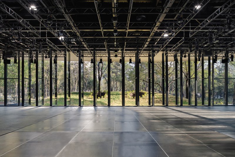 Interior view of exposed black structural grid ceiling and floor-to-ceiling glass overlooking trees