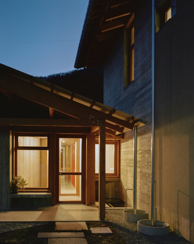 Illuminated timber entry pergola with exposed joists connecting concrete and plaster volumes at dusk