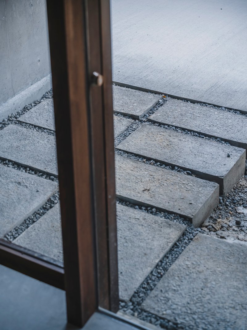 Detail of bronze door frame meeting concrete paving slabs set in gravel at threshold