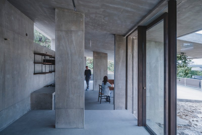 Two figures inhabit an open concrete interior with freestanding columns and a wall-mounted shelving unit