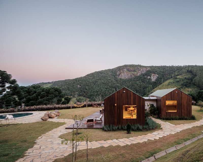 Two gabled timber volumes with illuminated interiors on flagstone paths at dusk beneath a mountain ridge