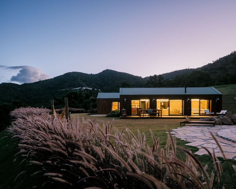 Low gabled structure with metal roof and glazed openings across a lawn with ornamental grasses at twilight