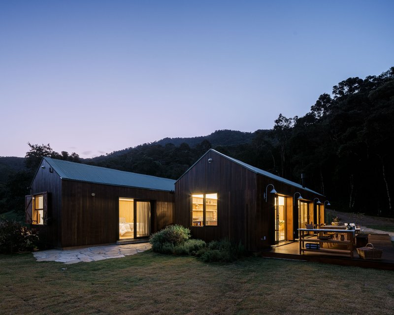 Timber-clad volumes with metal roofs and lit windows connected by a timber deck at evening