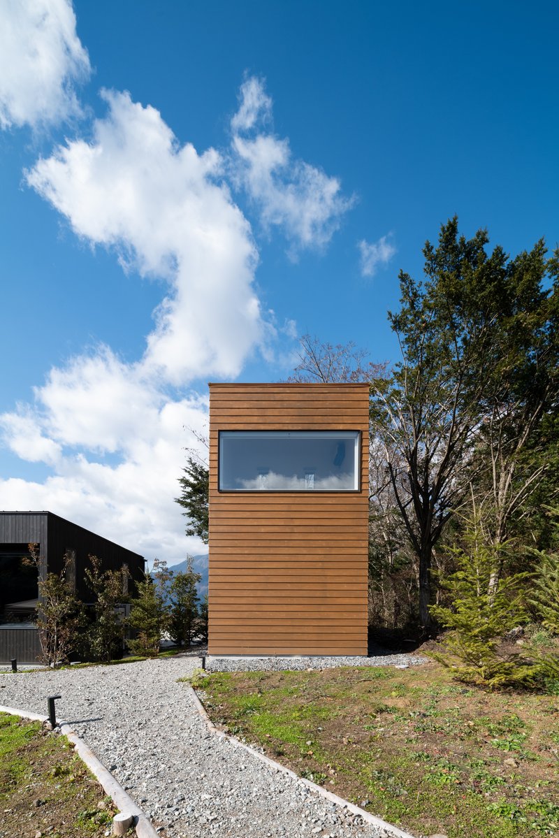 Freestanding cedar-clad tower with horizontal window band set on a gravel base beneath scattered clouds