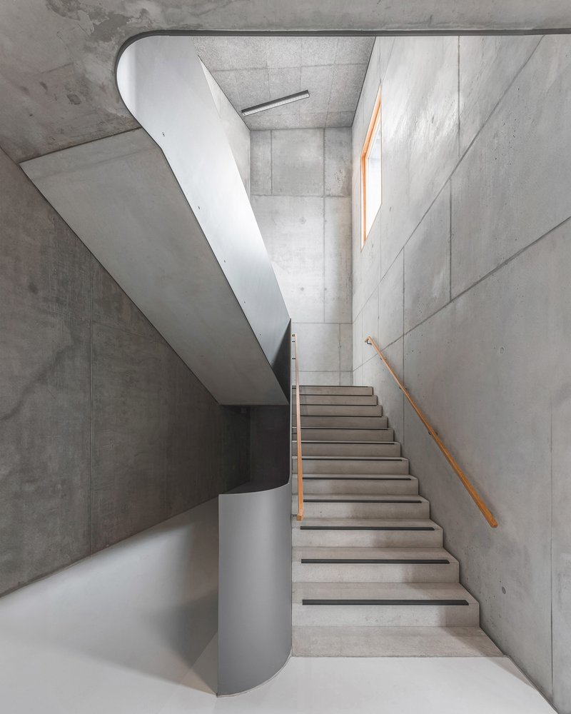 Concrete stairwell with curving white balustrade and timber handrail under natural skylight