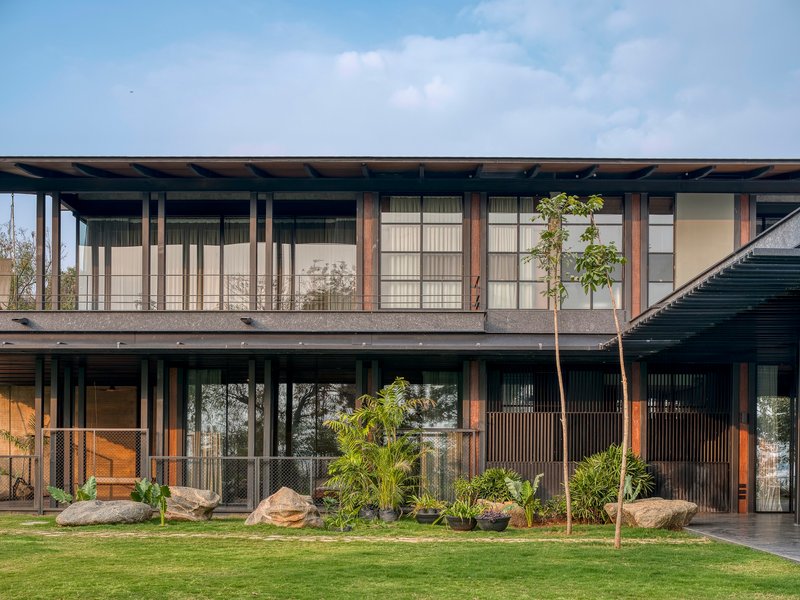 Two-story facade with vertical timber columns and glazing behind a lawn planted with palm and rock outcrops