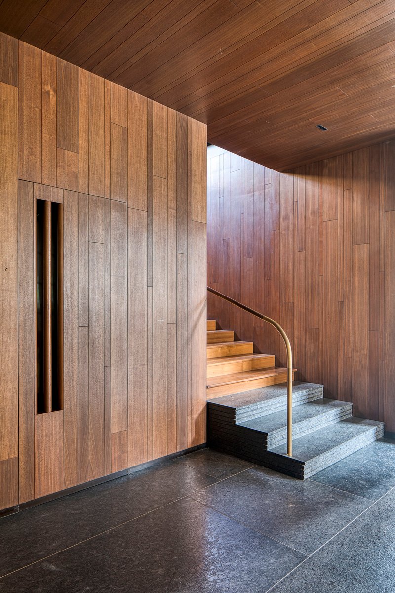 Interior staircase with timber treads and brass rail ascending beside a timber-clad wall with slot window