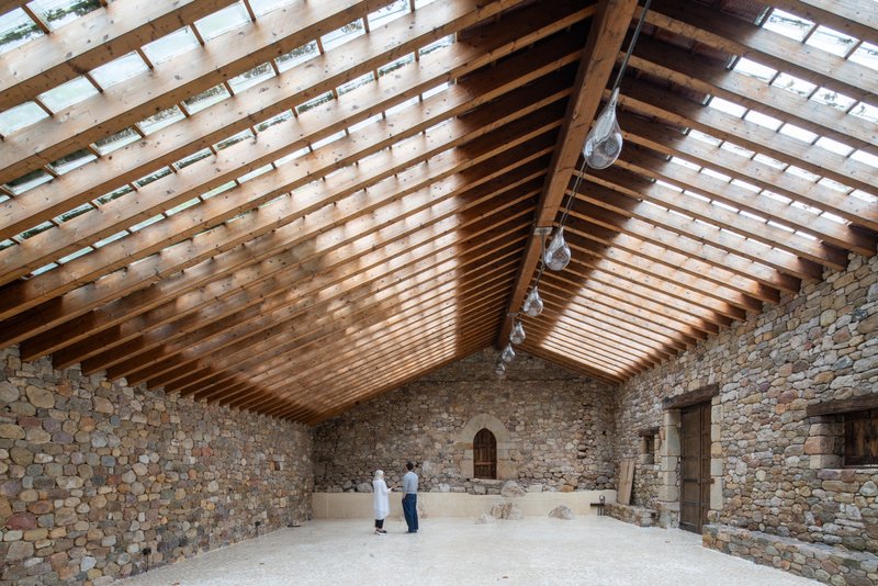 Gabled timber roof with skylight slats above a stone-walled interior hall with two visitors standing