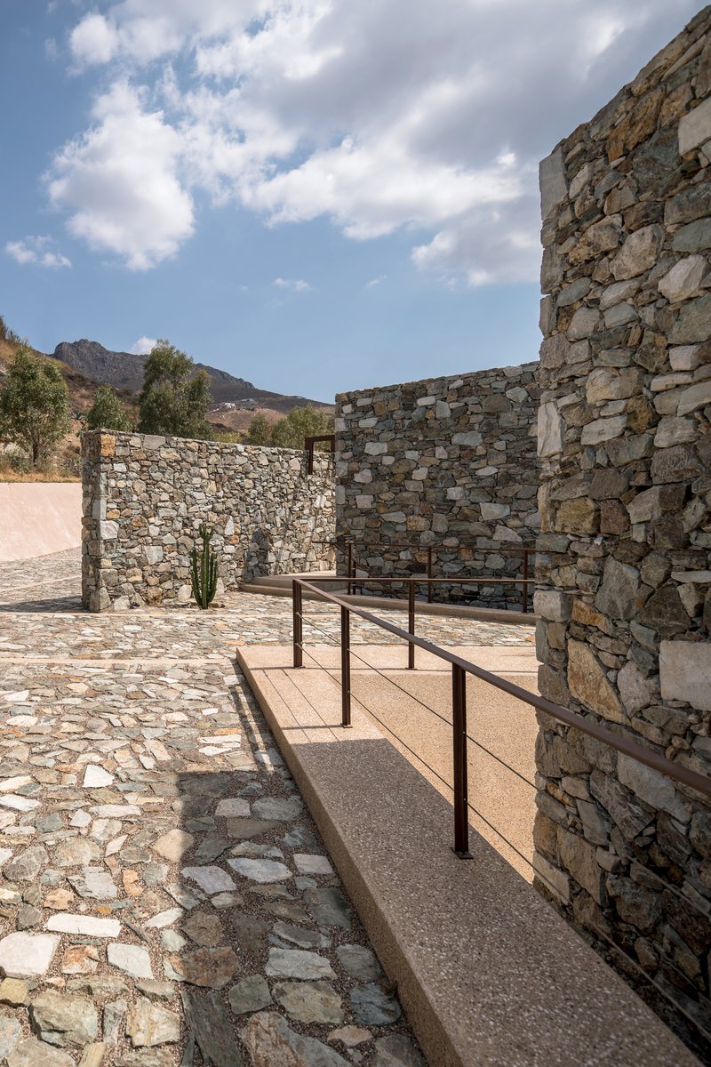 Stone-paved courtyard with metal-railed ramp between textured masonry walls under afternoon clouds