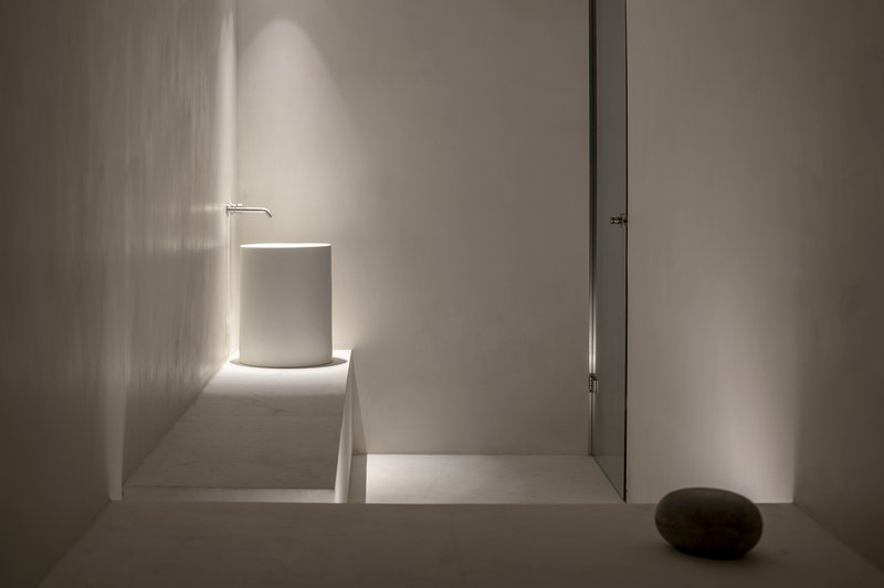 Minimalist corridor with backlit cylindrical volume and narrow slot of light along white plaster walls