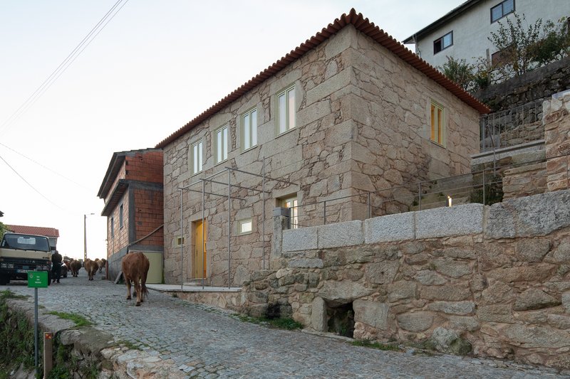 Two-story granite house with pale green windows along a cobblestone street with a passing cow