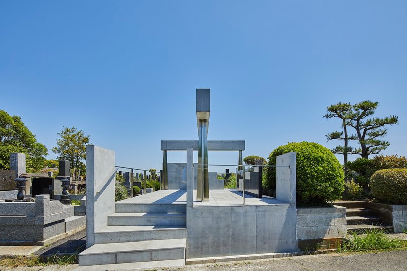 Concrete memorial platform with vertical columns supporting horizontal beams framing a glass monument under clear blue sky